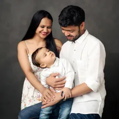 A classic family portrait against a simple, dark backdrop. The focus is entirely on the connection and love between the parents and their adorable little one.