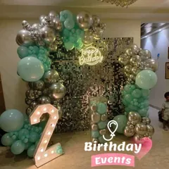 This modern second birthday decoration uses a unique color palette of mint green and silver. The design is centered around a sparkling shimmer wall backdrop, a neon sign, and a large light-up number '2'.