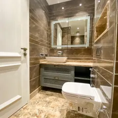 This bathroom combines functionality with classic design. It features a custom grey vanity with lattice-work doors, a large mirror with integrated lighting, and beautiful brown Italian marble on the floor and walls.