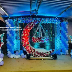 A magical fairy-themed naming ceremony stage. The design includes a floral crescent moon with a hanging cradle, set against a green backdrop with blue balloon pillars and fairy cutouts.