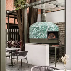 A traditional pizza oven clad in green and white mosaic chips stands as a playful focal point at Kana Cafe. This feature adds a unique, artisanal touch to the outdoor dining experience.