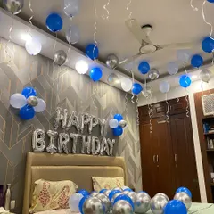 A simple yet stylish birthday room decoration. This setup includes blue, white, and silver chrome balloons on the ceiling and floor, with large "Happy Birthday" foil letters above the bed.