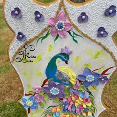 A close-up of a sculpted peacock, focusing on the details of its face and the surrounding purple flowers with pearl centers.