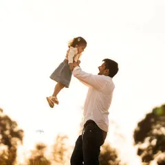 A joyful father lifts his daughter high in the air against a bright, sunlit sky during an outdoor family photoshoot.