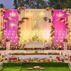 A fairytale engagement setup with a heart-shaped floral arch and monogram. The stage is bathed in warm light from hanging chandeliers and draped in pink and white fabrics, creating a romantic focal point.