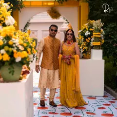 The couple poses at their Haldi ceremony. The decor features a custom-printed floral carpet and an entrance flanked by large arrangements of yellow and white flowers, creating a picture-perfect setting.