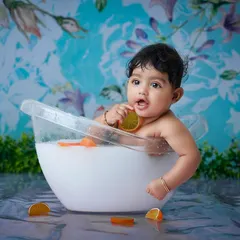 A fun and unique toddler "milk bath" session. The baby sits in a small tub with orange slices against a vibrant blue floral background, creating a playful and artistic image.