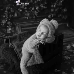A sleeping baby in a bear costume, posed on a miniature bed. The black and white edit gives this cute setup a classic, storybook feel.