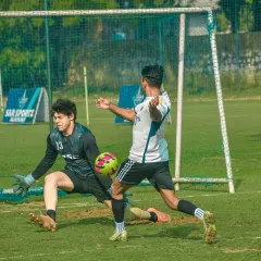 A goalkeeper makes a brave save at the feet of an attacker in a corporate match.