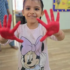 Getting a little messy is part of the fun and learning process here. This happy smile, framed by red-painted hands, captures the pure delight of our sensory art activities that help develop fine motor skills.