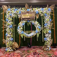 We also design naming ceremony stages using beautiful artificial flowers. This setup features a square floral arch with blue and white flowers, golden palm leaves, and a traditional wooden nameplate.