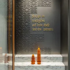 A close-up of a modern mandir design featuring the Gayatri Mantra in elegant gold lettering against a dark, intricately patterned wall. The marble steps and decorative pagodas create a serene and sacred focal point.