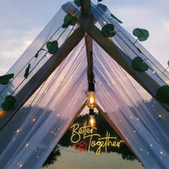 A romantic and intimate anniversary celebration setup in a tent by a lake in Karjat. The warm glow of the 'Better Together' neon sign and fairy lights creates a cozy, unforgettable atmosphere at dusk.