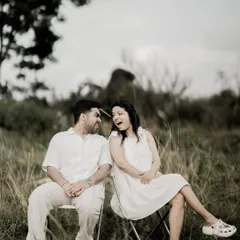 A black and white shot of a couple sitting on chairs in a field, sharing a laugh, capturing a genuine and happy moment.
