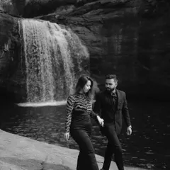 A dramatic black and white image of a couple walking by a waterfall, highlighting the contrast and texture of the natural landscape.