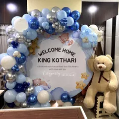 A 'Welcome Home' decoration for a baby boy with a classic teddy bear theme. The setup includes a custom circular backdrop, a blue, white, and silver balloon arch, 'BABY' blocks, and a large teddy bear prop.