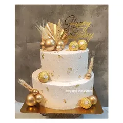 A luxurious two-tier cake featuring a textured white finish and gold accents. It's decorated with Ferrero Rocher chocolates, gold spheres, and dried wheat for a modern and celebratory look.
