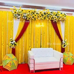 A classic Haldi or Mehendi setup with a bright yellow backdrop, marigold garlands, and a pop of red and white draping. The 'Mehendi' neon sign and traditional umbrellas add to the festive charm.