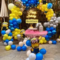 A playful Minions themed birthday party. The outdoor setup includes a blue and yellow balloon arch, multiple Minion character figures, and a 'Happy Birthday' neon sign.