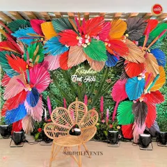 For a 'First Holi' celebration, we created this explosion of color. A lush green wall serves as the base for vibrant, multi-colored dried palm leaves, creating a joyful and energetic photo backdrop. The natural rattan peacock chair adds a touch of bohemian chic to the setup.