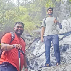 Two trekkers posing in front of a waterfall, fully equipped with backpacks and trekking poles.