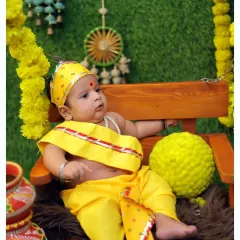 An alert baby dressed in a yellow Krishna costume sits on a miniature swing decorated with marigold garlands. This setup creates a playful and festive atmosphere for the photoshoot.