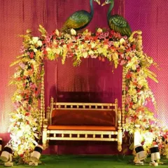 A beautiful jhula for a Janmashtami or Teej celebration. The swing is covered in a dense arrangement of colorful flowers, with two peacock statues sitting on top, creating a festive and traditional look.