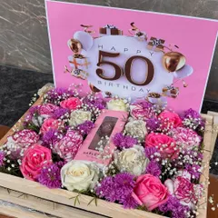 A beautiful floral crate celebrating a 50th birthday. The mix of pink, purple, and white flowers creates a vibrant and joyful arrangement, with a chocolate bar nestled in the center.
