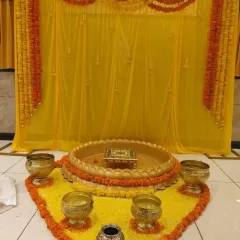 A simple and traditional Haldi ritual area. The space is defined by a yellow floral carpet, brass bowls, and a backdrop of yellow drapes and marigold garlands.