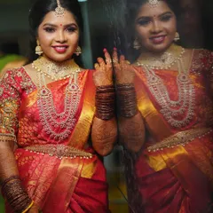 A stunning reception look featuring a vibrant red saree and layered diamond necklaces. The makeup is glamorous, with a bright lip and defined eyes that sparkle next to her reflection.