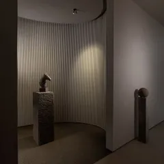 Inside a lighting showroom, a curved, textured wall becomes a canvas for a play of light and shadow, with a single spotlight dramatically highlighting a sculpture on a stone pedestal.