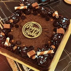 A close-up of my decadent Rakhi brownie cake. It's a rich, fudgy brownie base, generously decorated with chocolate frosting, chocolate chunks, and wafer rolls, complete with a 'Happy Rakhi' topper.