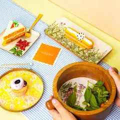 A lifestyle shot featuring hands holding a wooden bowl, with colorful, artisanal plates and pastries creating a vibrant table scene.