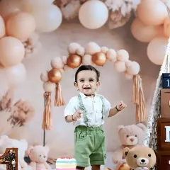 A happy toddler celebrates his birthday with a boho-themed setup, complete with teddy bears, pampas grass, and a colorful cake.