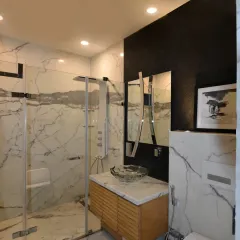 The bathroom in Gupta Residence features a luxurious modern design with white marble-like surfaces, a contrasting black wall, and a floating wooden vanity with a glass bowl basin. A sculptural mirror and enclosed glass shower enhance its elegance.