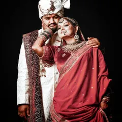 A stylish and confident wedding portrait. The bride playfully holds her groom's face, showcasing her intricate henna and jewelry, while he looks on with affection. The dark background makes their vibrant attire pop.