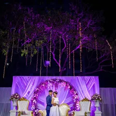 A grand reception stage with the couple framed by an arch of purple flowers and hanging lights. This portrait showcases the beautiful decor and scale of the event.