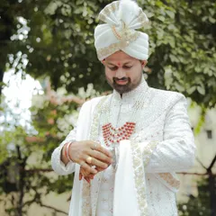 A full-length outdoor portrait of the groom adjusting his attire. The natural light and green background provide a fresh contrast to his elegant white wedding sherwani.