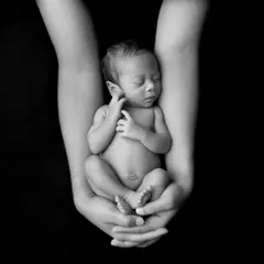 A powerful black and white fine art photograph showing a sleeping newborn held securely in their parent's cupped hands against a stark black background, emphasizing their tiny, delicate form.