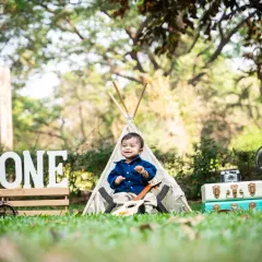 A beautifully styled outdoor first birthday photoshoot featuring a baby boy sitting in front of a teepee, surrounded by props like a 'ONE' sign, vintage suitcases, and a globe.