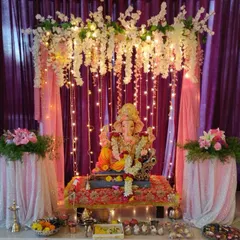 A dreamy Ganpati mandap created with a canopy of white flowers and cascading fairy lights against a purple curtain. The sequined fabric on the side tables adds a soft, celebratory shimmer.