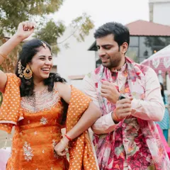 Pure happiness at a sundowner mehendi. The couple is dancing, and their coordinated floral and pastel outfits look fantastic. The groom's floral jacket and the bride's orange sharara are a perfect match.