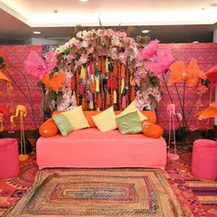 The full seating area for the Mehendi, designed with a boho-chic vibe. The setup includes a colorful tassel backdrop, large paper flowers, flamingo props, and decorated umbrellas.