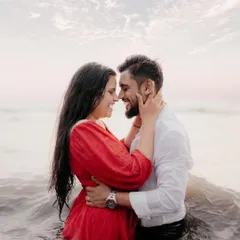 A passionate and playful shot of a couple embracing in the ocean waves, laughing together. The woman is wearing a striking red dress, creating a beautiful contrast with the water for a memorable pre-wedding photo.