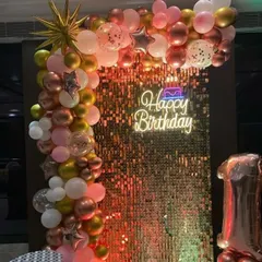 A birthday party with a rose gold shimmer wall, a 'Happy Birthday' neon sign, and a balloon garland.