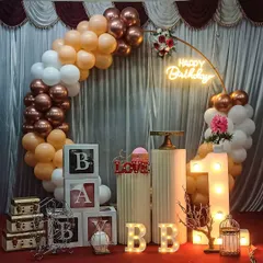 A chic and modern setup featuring a circular frame adorned with peach, white, and rose gold balloons. The scene is completed with 'BABY' blocks, light-up marquee letters, and a pedestal for the cake, perfect for a first birthday or baby shower.