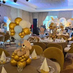 An elegant table centerpiece for a teddy bear-themed party, featuring a teddy bear holding a bouquet of gold, white, and pastel blue balloons.