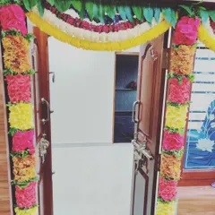 A beautifully decorated doorway for a housewarming event. The door frame is lined with colorful flower arrangements and a traditional mango leaf toran at the top, signifying prosperity and good fortune.