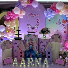 A magical butterfly-themed party in shades of pink and purple. The decoration includes themed pedestals, large butterfly wing props, house-shaped displays, and light-up letters for the birthday girl's name.