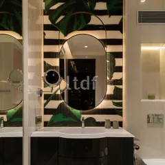 This bathroom vanity evokes a sense of luxury with premium materials and warm, integrated lighting. The elliptical mirror with its sleek frame is set against a bold backdrop of botanical prints and stripes, adding refinement and creating a serene retreat.